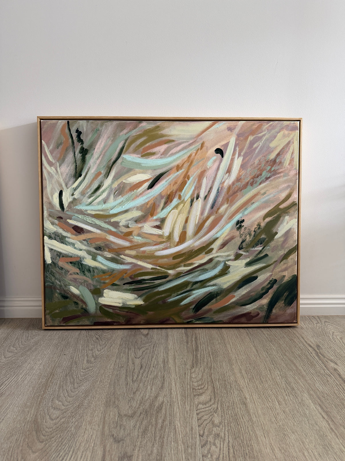 An abstract framed painting with sweeping brushstrokes in beige, brown, white, green, and blue-gray tones, leaning against a white wall on a light wood floor.