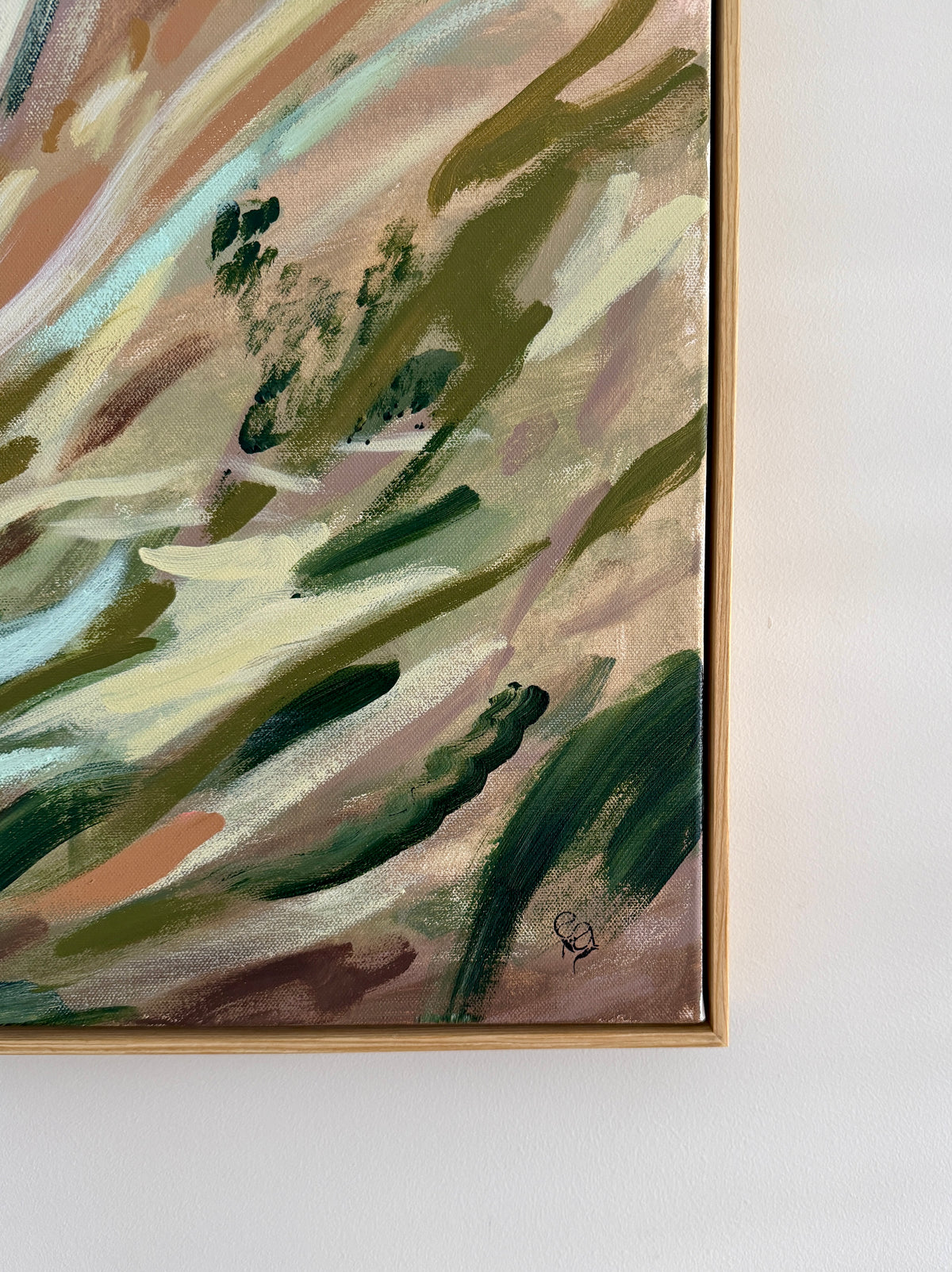 An abstract framed painting with sweeping brushstrokes in green, beige, brown, and soft peach tones, shown hanging against a white wall.