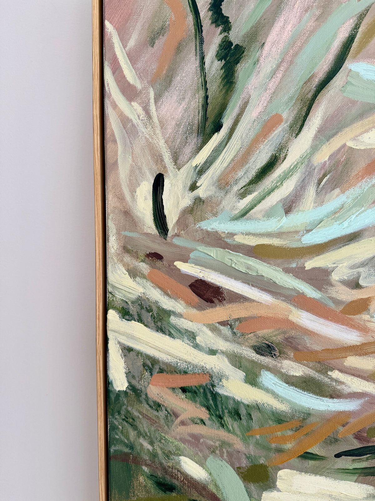 Abstract framed painting with expressive brushstrokes in soft green, cream, blush pink, tan, and light blue, shown hanging against a light gray wall.