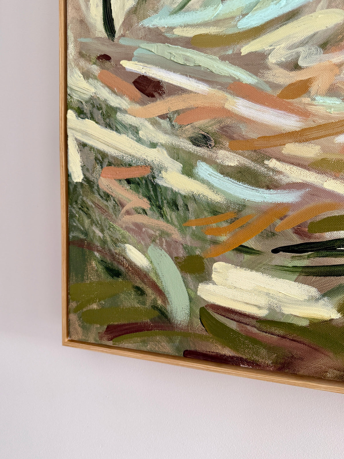 An abstract framed painting with expressive brushstrokes in olive green, cream, tan, light blue, and brown, shown hanging on a white wall.