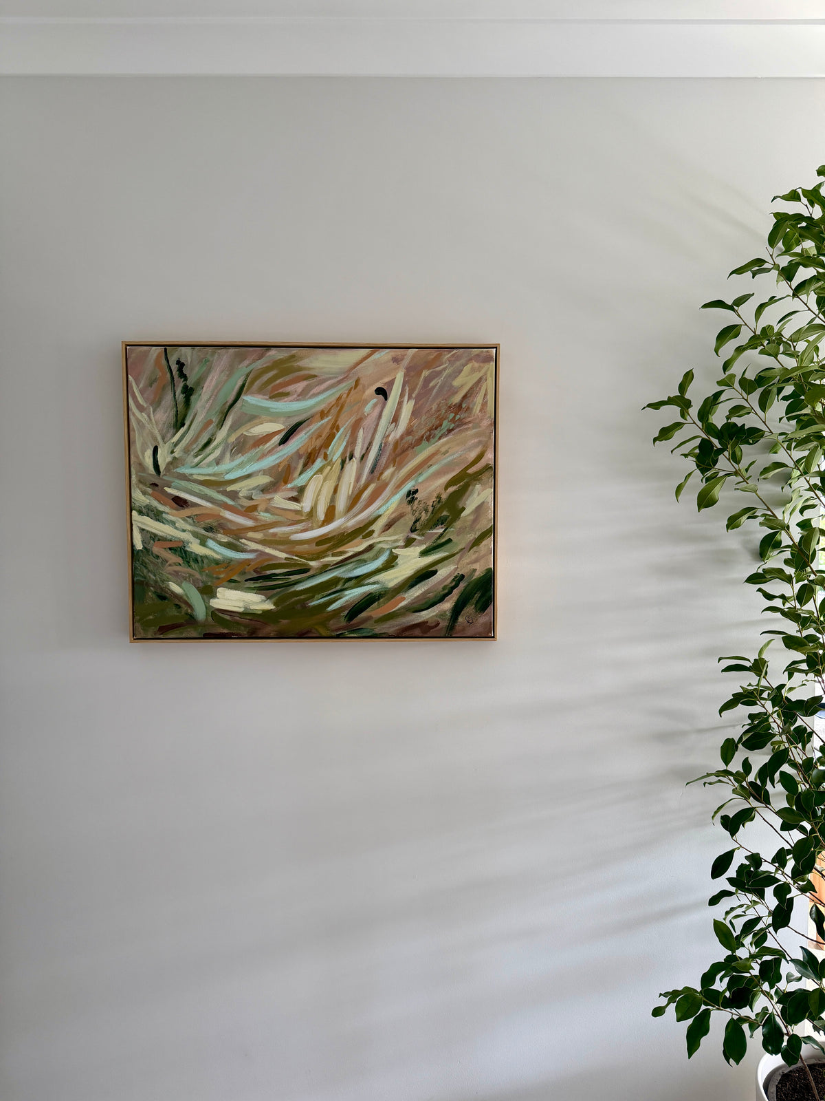 An abstract framed painting with sweeping brushstrokes in beige, green, brown, and cream tones, displayed on a white wall next to a leafy green plant.