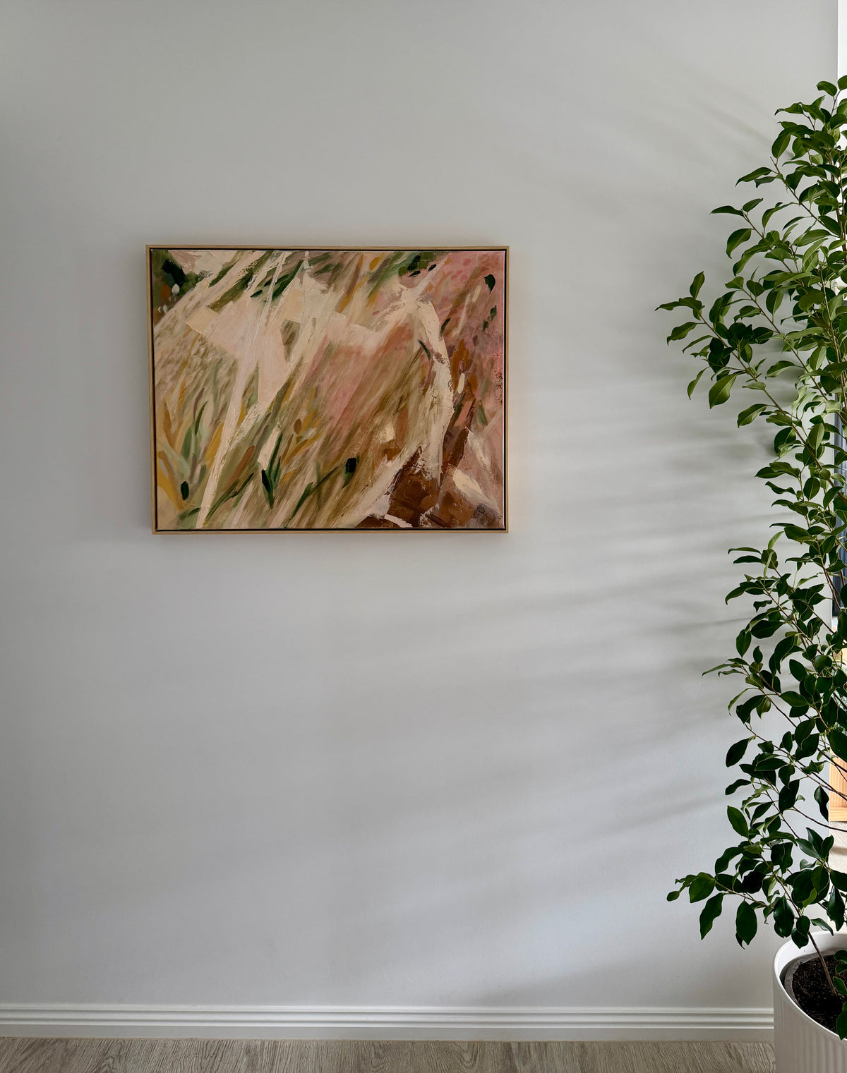 An abstract framed painting with earthy tones of beige, brown, pink, and green hangs on a light gray wall next to a green potted plant.